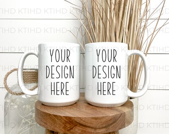 Puede incluir: Dos tazas de cerámica blanca con texto negro que dice "Your Design Here". Las tazas están sobre una superficie de madera con un jarrón de flores secas en el fondo.