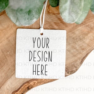 May include: A white square tag with the text "YOUR DESIGN HERE" in black, hanging by a white string. The tag rests on a wooden surface with green leaves in the background.