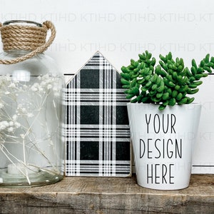 May include: A white ceramic planter with the text "YOUR DESIGN HERE" in black lettering. The planter is filled with a green succulent plant and is sitting on a wooden shelf next to a clear glass jar with dried white flowers and a black and white plaid house-shaped decor.