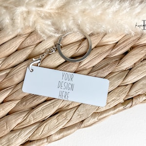 May include: A white rectangular keychain with the text "YOUR DESIGN HERE" on a woven straw background. The keychain has a silver ring and chain.