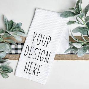 May include: White kitchen towel with the text "YOUR DESIGN HERE" printed in black. The towel is lying on a brown and black checkered ribbon with green leafy sprigs on either side.