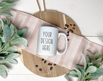 May include: A white ceramic mug with a handle on the right side, featuring the black text "YOUR DESIGN HERE" printed in a simple sans-serif font on the front surface. The mug is positioned centrally, surrounded by green leaves and coffee beans.