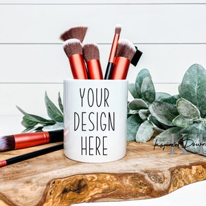 Puede incluir: Una taza de cerámica blanca con el texto "YOUR DESIGN HERE" contiene varios pinceles de maquillaje con mangos naranjas y cerdas marrones. La taza está sobre una tabla de madera con vegetación.