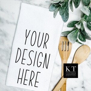 May include: White kitchen towel with the text "YOUR DESIGN HERE" printed in black. Two wooden spoons and a small black rectangle with the text "KT INSPIRED DOWNLOADS" are on a white marble surface.