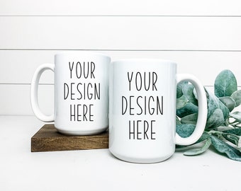 Puede incluir: Dos tazas de cerámica blanca con texto negro que dice "Your Design Here" sobre una superficie de madera blanca con un posavasos de madera marrón. Un brote de follaje verde está en el fondo.
