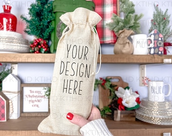 May include: A beige burlap bag with a drawstring closure. The bag is held in a hand and has the text "YOUR DESIGN HERE" printed on the front. The bag is in front of a shelf with various Christmas decorations.