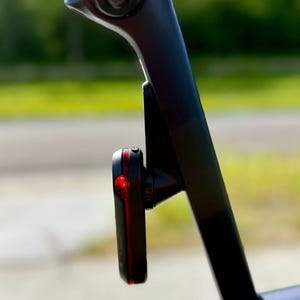 May include: A black bicycle tail light with a red light is mounted on the seat post of a bicycle. The light is attached to a black bracket. The background is blurred, showing a green area and a road.