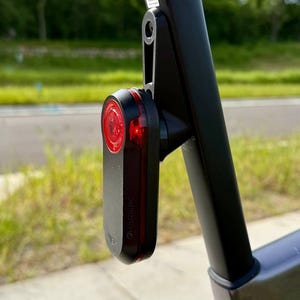 May include: A black Garmin bike tail light with a red lens is mounted on a black bike frame. The light is rectangular with rounded edges and has the Garmin logo on the side. The bike frame is also black.