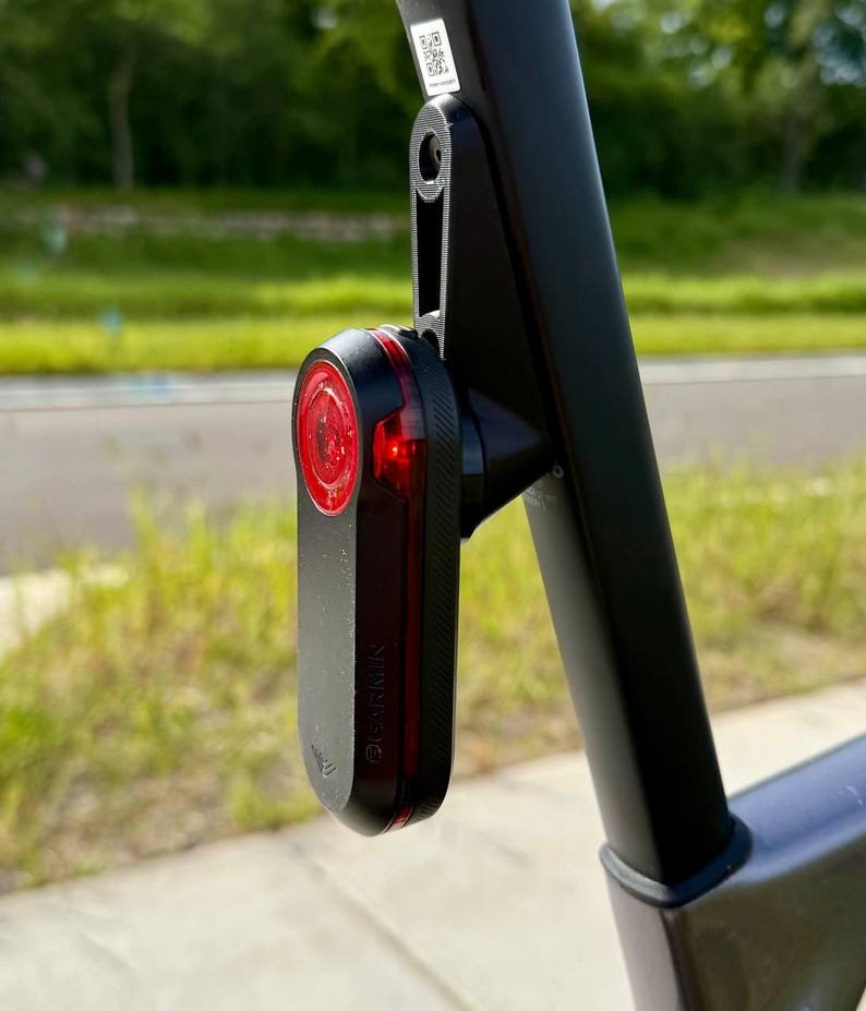 May include: A black Garmin bike tail light with a red lens is mounted on a black bike frame. The light is rectangular with rounded edges and has the Garmin logo on the side. The bike frame is also black.
