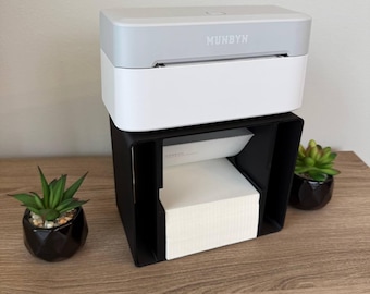 May include: A white and gray label printer with the brand name "MUNBYN" printed on the front. The printer sits atop a black label holder containing a stack of white labels. Two small potted succulents are on the wooden surface.