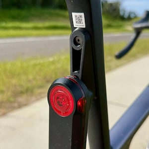 May include: Black Garmin bike tail light attached to a bicycle seat post. The light has a red lens and a black body. The seat post is black and has a QR code sticker.