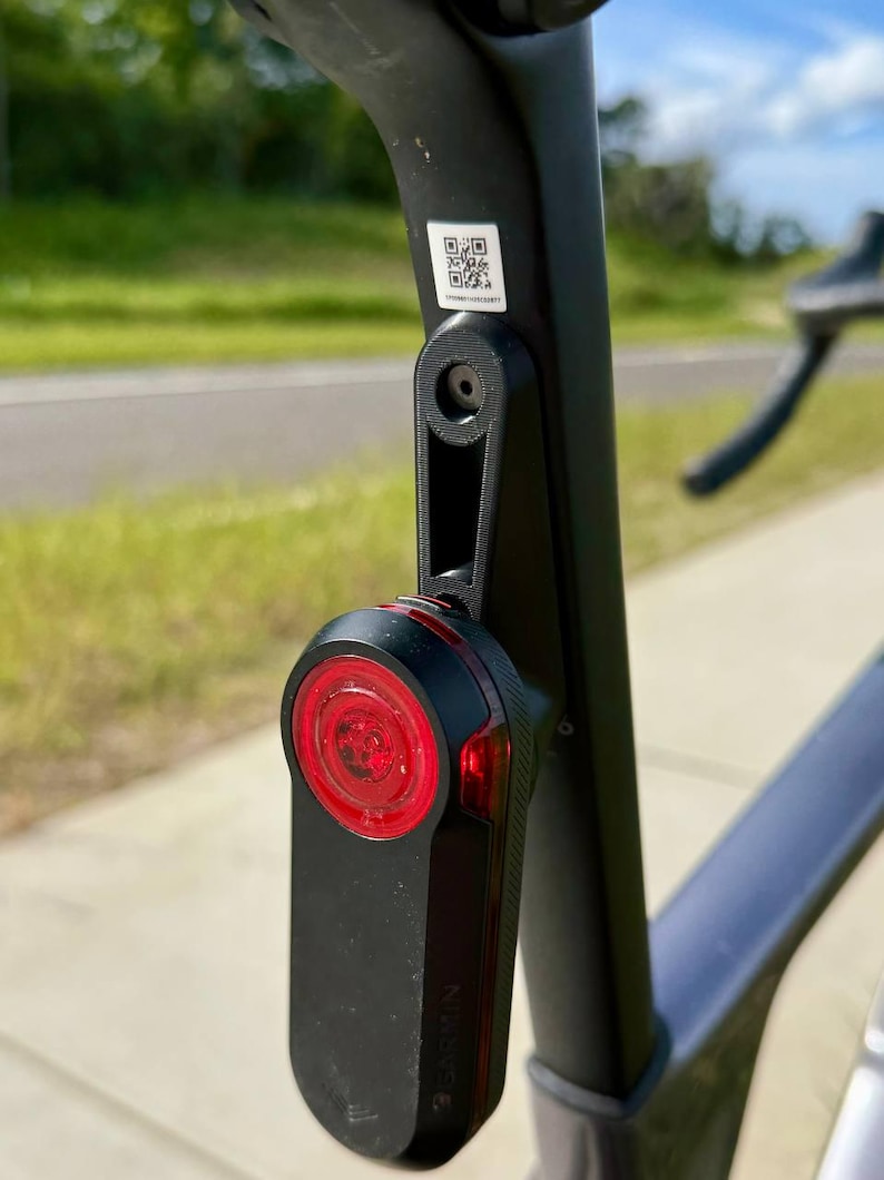May include: Black Garmin bike tail light attached to a bicycle seat post. The light has a red lens and a black body. The seat post is black and has a QR code sticker.