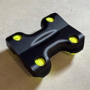 May include: A black, T-shaped device holding four yellow tennis balls. The device has rounded edges and is designed to hold the balls securely. The tennis balls are bright yellow, contrasting with the black holder. The item appears to be made of a solid material.