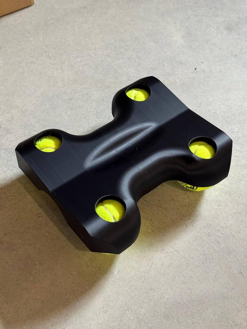 May include: A black, T-shaped device holding four yellow tennis balls. The device has rounded edges and is designed to hold the balls securely. The tennis balls are bright yellow, contrasting with the black holder. The item appears to be made of a solid material.