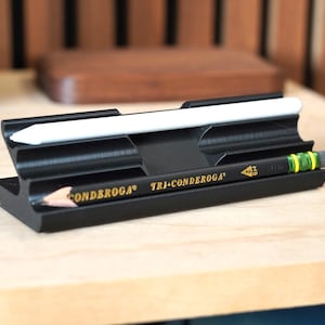 May include: A black desk organizer holds a white stylus and a yellow and black pencil. The pencil is a Tri-Conderoga HB. The organizer is made of a solid black material and sits on a light wood surface.