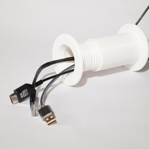 May include: A white cable management grommet with multiple cables extending from it. Cables include a USB, HDMI, and Ethernet cable. The grommet is cylindrical with a flared edge on each end.