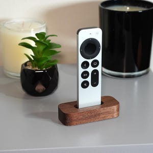 May include: A white remote control stands in a wooden holder on a gray surface. A small potted plant and a candle in a glass jar are in the background. The remote has black buttons.