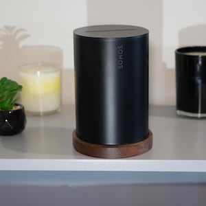 May include: A black cylindrical speaker with the brand name "SONOS" on the side, resting on a wooden base. The speaker is on a gray surface, accompanied by a small potted plant and two candles in glass containers.