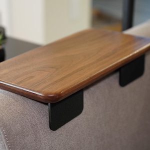 May include: A rectangular wooden sofa arm tray with a smooth, polished brown grain finish. The tray features two black metal side supports that extend downward to grip the sides of a gray upholstered sofa arm, providing a flat surface.