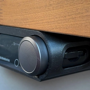 May include: A black SteelSeries audio device with a large, round volume control knob. The device is mounted under a wooden surface and held in place by a black bracket. The SteelSeries logo is visible on the device.