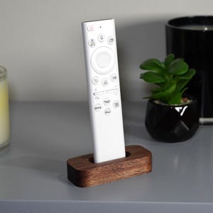 May include: A white remote control is placed in a dark brown wooden holder. The remote has multiple buttons and a circular control pad. A candle and a small potted plant are in the background.