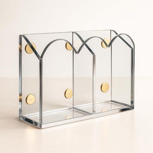 May include: Clear acrylic desk organiser with four arched compartments. The organiser has a rectangular base and is accented with gold-coloured circular fasteners. The design is modern and minimalist, suitable for office or home use.