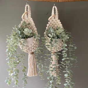 Op de afbeelding: Twee macramé plantenhangers met houten ringen, elk met een kleine witte keramische pot met een groene, bladerrijke plant. De planten hangen aan een houten stok tegen een groene muur.