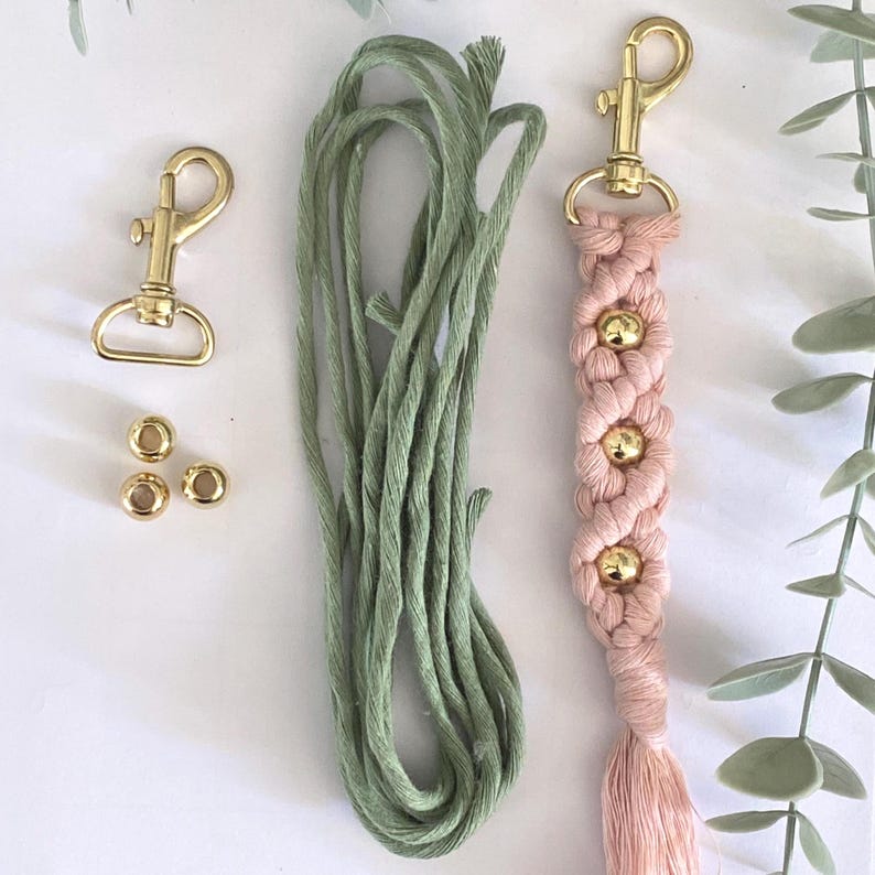 May include: A macrame keychain kit featuring a pink woven design with gold beads and a tassel. The kit includes a gold-colored clasp, gold beads, and green cord, all arranged on a white surface. The keychain is approximately 6 inches long.