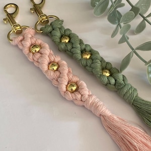 K&ouml;nnte beinhalten: Zwei Makramee-Schl&uuml;sselanh&auml;nger, einer rosa und einer gr&uuml;n, mit goldfarbener Hardware. Jeder Schl&uuml;sselanh&auml;nger hat ein geflochtenes Design mit goldenen Perlen und einem Quastenanh&auml;nger. Die Schl&uuml;sselanh&auml;nger werden auf einer wei&szlig;en Oberfl&auml;che mit gr&uuml;nem Laub im Hintergrund pr&auml;sentiert.