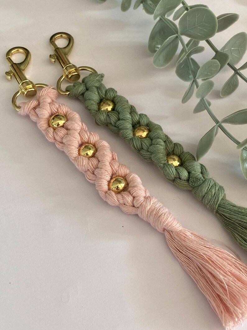 K&ouml;nnte beinhalten: Zwei Makramee-Schl&uuml;sselanh&auml;nger, einer rosa und einer gr&uuml;n, mit goldfarbener Hardware. Jeder Schl&uuml;sselanh&auml;nger hat ein geflochtenes Design mit goldenen Perlen und einem Quastenanh&auml;nger. Die Schl&uuml;sselanh&auml;nger werden auf einer wei&szlig;en Oberfl&auml;che mit gr&uuml;nem Laub im Hintergrund pr&auml;sentiert.