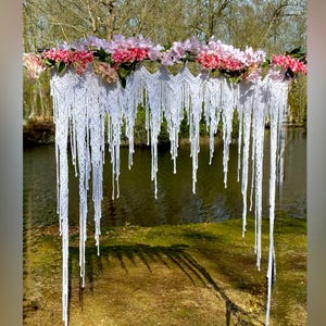 May include: White macrame wedding arch with pink flowers hanging from it. The arch is set up outdoors in front of a body of water.