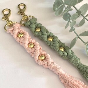 May include: Two macrame keychains, one pink and one green, with gold-colored hardware and decorative gold beads. The keychains have a braided design and a tassel at the end. The background is white with a hint of greenery.