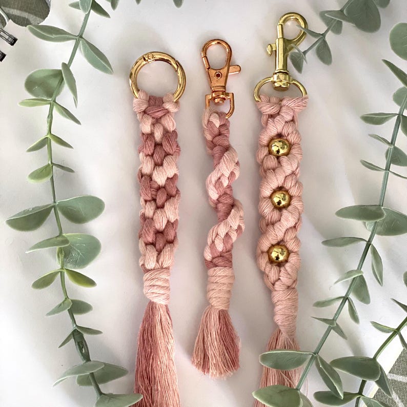 May include: Three macrame keychains in shades of pink and cream, each with a different design. The keychains have gold or rose gold hardware and include tassels. The keychains are displayed on a white surface with green foliage.