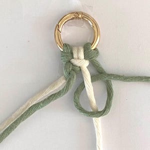 K&ouml;nnte beinhalten: Nahaufnahme eines Makramee-Schl&uuml;sselanh&auml;ngers mit einem goldfarbenen Verschluss. Der Makramee-Knoten weist gr&uuml;ne und cremefarbene Kordeln auf, die ein strukturiertes und handgefertigtes Aussehen erzeugen. Das Design zeigt eine Kombination aus Farbe und Textur.