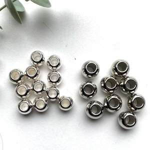 May include: A group of silver metal beads with a hole in the centre. The beads are arranged in two piles on a white surface.