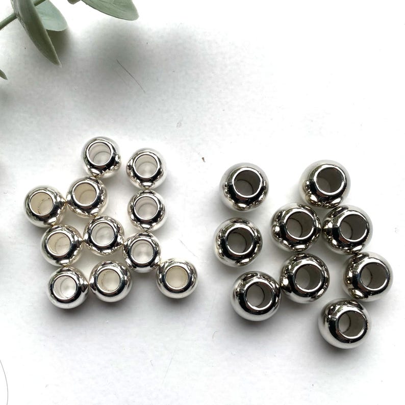 May include: A group of silver metal beads with a hole in the centre. The beads are arranged in two piles on a white surface.