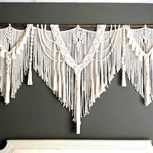 Op de afbeelding: Een grote witte macramé wandhanger met een houten stok aan de bovenkant. De macramé is gemaakt van verschillende knopen en patronen en heeft een bohemian stijl.