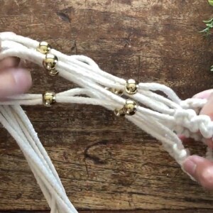 May include: Close-up of a white macrame plant hanger with gold beads. The macrame is being tied together with the beads in the center of the image.