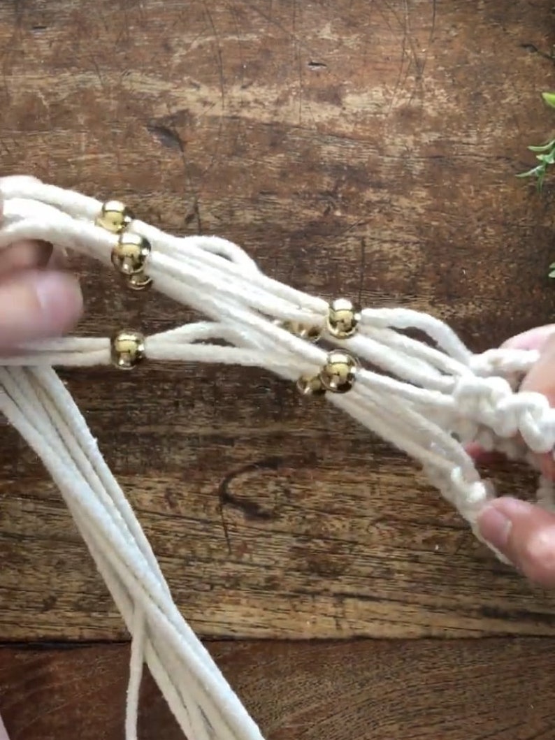 May include: Close-up of a white macrame plant hanger with gold beads. The macrame is being tied together with the beads in the center of the image.