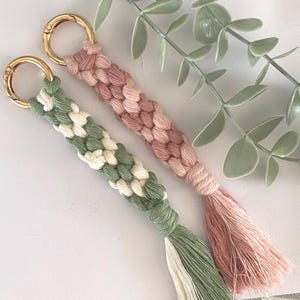 May include: Two macrame keychains with gold-colored clasps. One is green and white, the other is pink and peach. Both have braided designs and matching tassels. Decorative greenery is in the background.