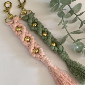 K&ouml;nnte beinhalten: Zwei Makramee-Schl&uuml;sselanh&auml;nger, einer in Hellrosa und der andere in Olivgr&uuml;n, jeweils mit einem Blumendesign und goldenen Perlen. Beide Schl&uuml;sselanh&auml;nger haben goldfarbene Verschl&uuml;sse und passende Quasten. Ein handgefertigtes Accessoire.