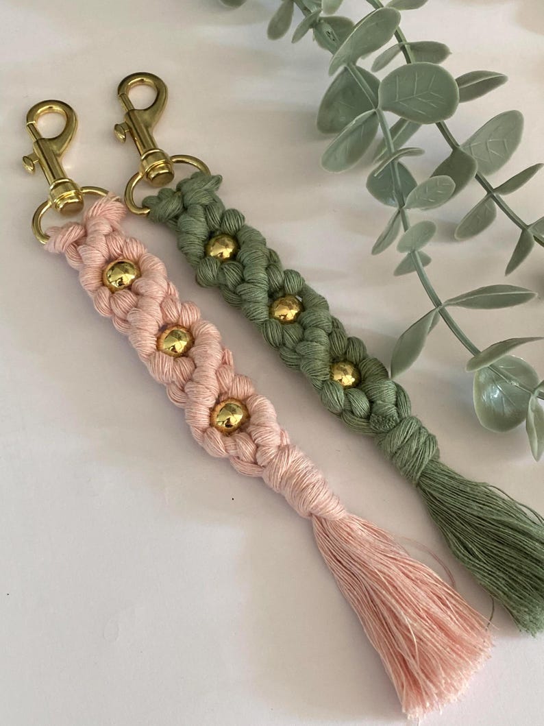 K&ouml;nnte beinhalten: Zwei Makramee-Schl&uuml;sselanh&auml;nger, einer in Hellrosa und der andere in Olivgr&uuml;n, jeweils mit einem Blumendesign und goldenen Perlen. Beide Schl&uuml;sselanh&auml;nger haben goldfarbene Verschl&uuml;sse und passende Quasten. Ein handgefertigtes Accessoire.