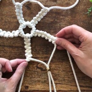 May include: A white macrame wall hanging with a gold bead at the bottom. The macrame is made with a knotting technique that creates a starburst pattern.