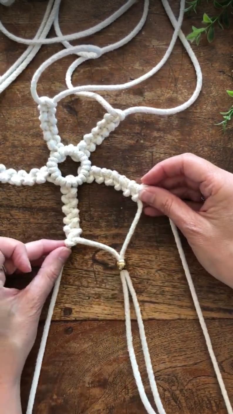 May include: A white macrame wall hanging with a gold bead at the bottom. The macrame is made with a knotting technique that creates a starburst pattern.
