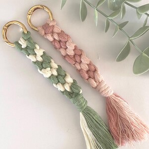 K&ouml;nnte beinhalten: Zwei handgefertigte Makramee-Schl&uuml;sselanh&auml;nger. Einer ist gr&uuml;n und wei&szlig;, der andere rosa. Beide haben goldfarbene Metallverschl&uuml;sse und passende Quasten. Die Schl&uuml;sselanh&auml;nger sind aus gewebter Baumwollschnur.