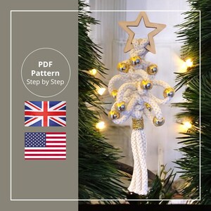Könnte beinhalten: Handgefertigter Makramee-Weihnachtsbaumschmuck. Der weiße Baum ist mit goldfarbenen Kugeln und einem Stern aus Holz verziert. Der Sockel hat ein goldenes Band und eine Quaste. Das Bild enthält den Text "PDF Pattern Step by Step".