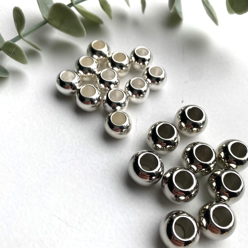 May include: A collection of silver-toned metal beads, some are round and some are slightly oval shaped. The beads are arranged in two groups on a white surface.