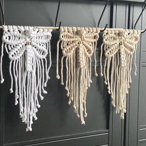 Op de afbeelding: Drie macramé wandkleden in wit en beige, elk met een vlinderachtig ontwerp. De wandkleden hebben ingewikkelde knoop patronen en lange, bungelende franjes. Ze zijn opgehangen aan een houten deuvel en versierd met kleine gouden kralen.