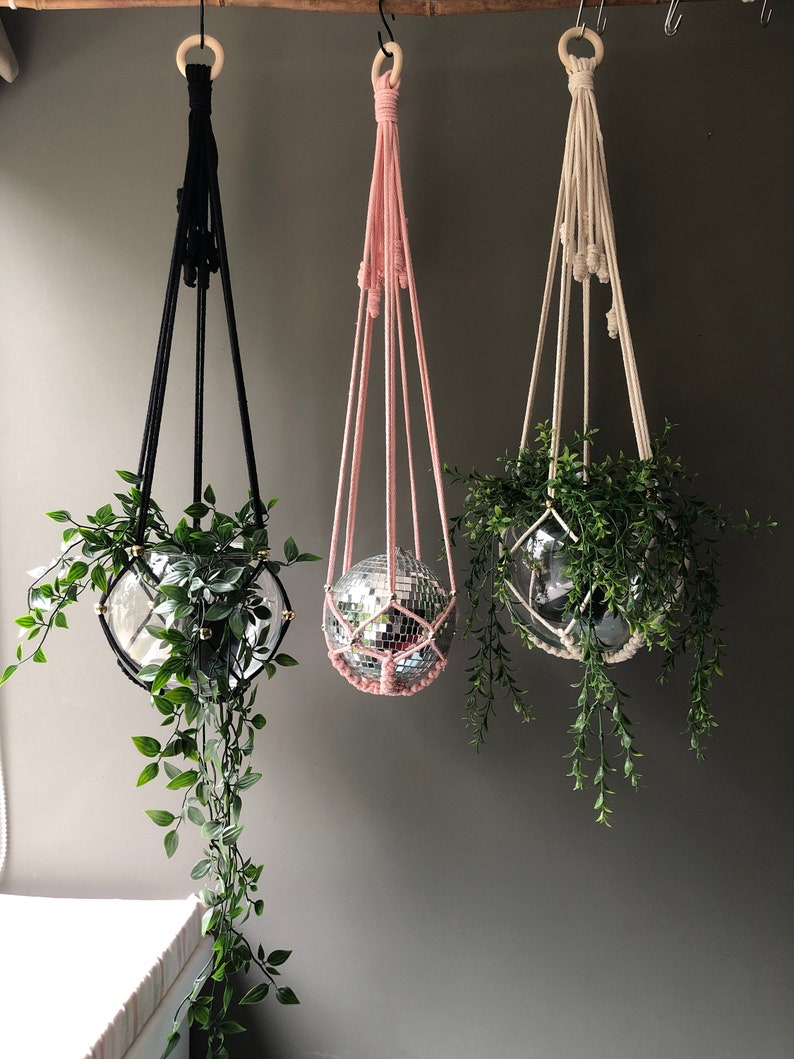 May include: Three macrame plant hangers with wooden rings, each holding a potted plant and a disco ball. The hangers are black, pink, and white.