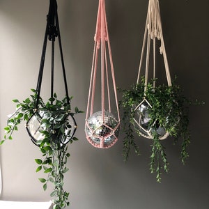Op de afbeelding: Drie macramé plantenhangers met houten ringen, elk met een potplant en een discobal. De hangers zijn zwart, roze en wit.